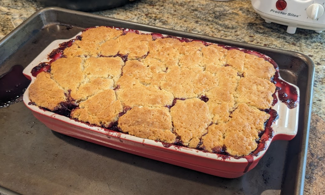 An overfilling baking dish on a sheetpan with pastry on top and blackberry filling leaking out. 