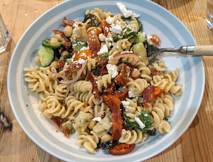 A blue bowl filled with dressed fusili noodles, olives, peppers, tomatoes, chickpeas, onion and cucumbers, topped with feta cheese. 