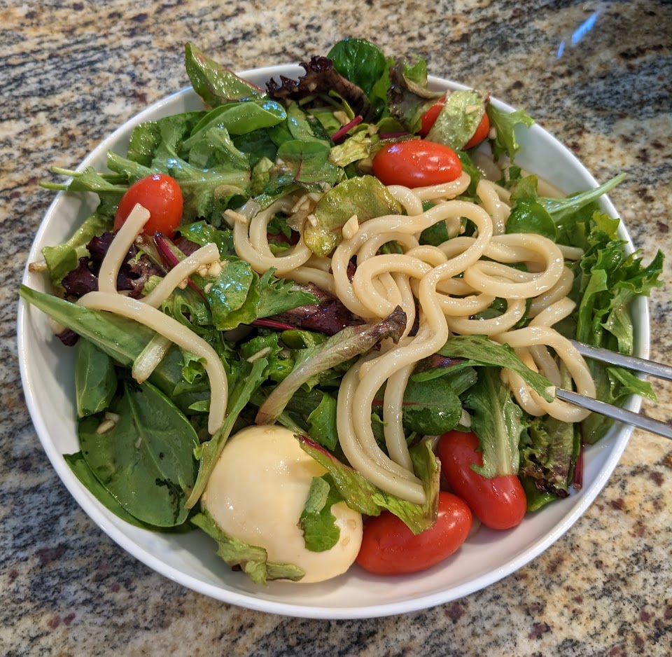 A generous portion of dressed udon noodles with small leafy greens, tomatoes, and a hard boiled egg in a shallow bowl.